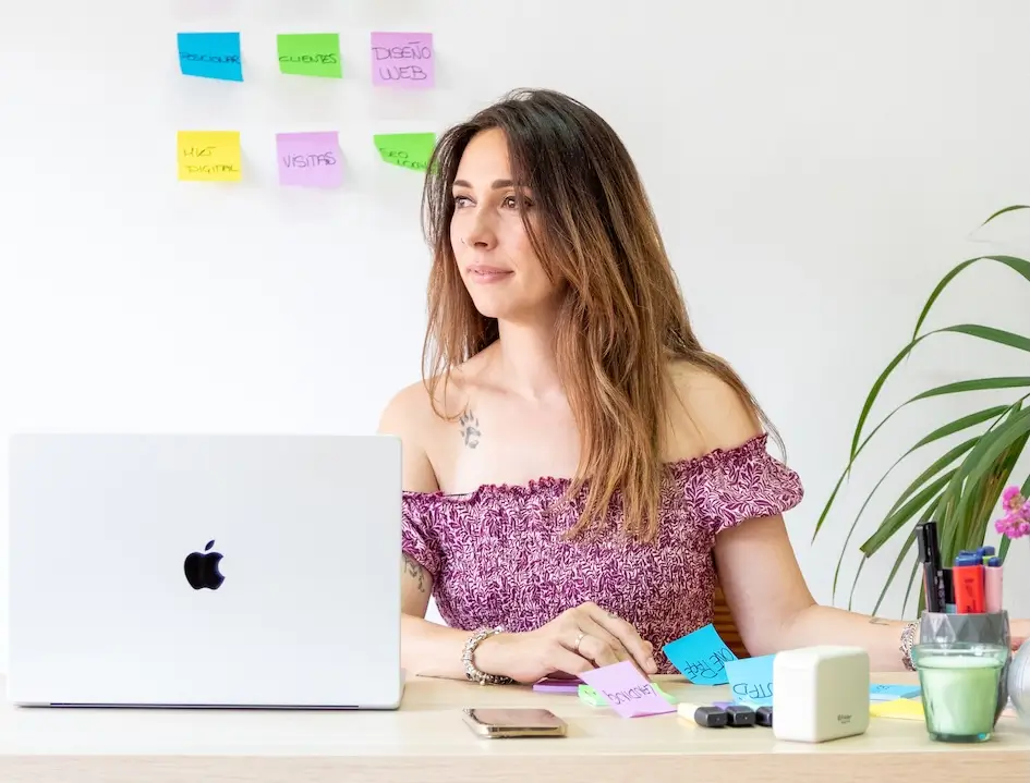 Mujer en escritorio con portátil, notas y planta, trabajando en oficina. Laura G.Arias.