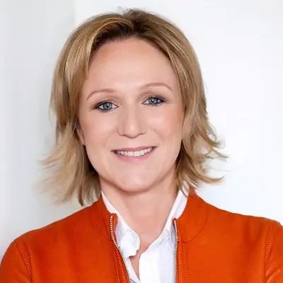 Rosa Gorriz con chaqueta naranja sobre camisa blanca sonríe frente a fondo claro.