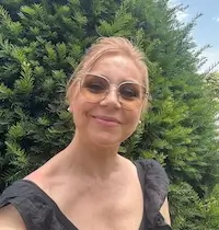 Mujer con gafas de sol y top negro sonríe al aire libre frente a arbustos verdes